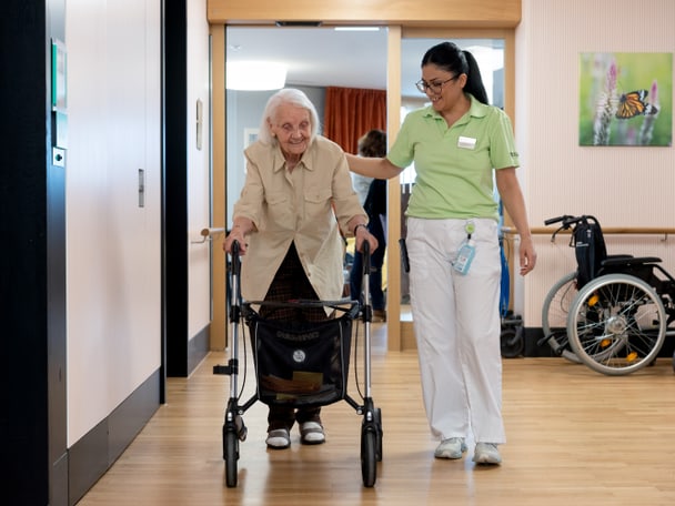 Eine Pflegefachperson begleitet einen älteren Menschen am Rollator.