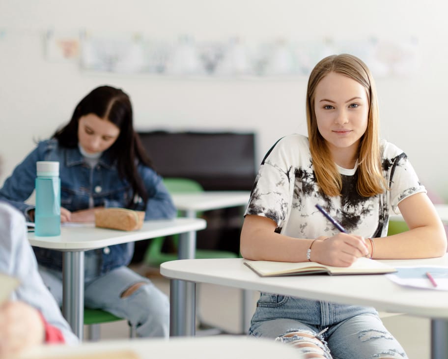 Schülerin sitzt im Klassenzimmer am Tisch und schaut in die Kamera.